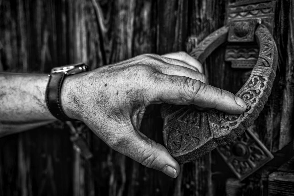 Detailed black and white photo of a hand gripping an intricately designed door knocker on a wooden gate.